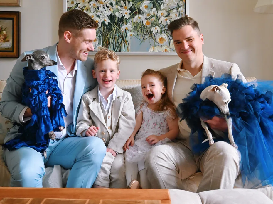 Two men, a young boy, and a girl sitting with two dogs in blue outfits